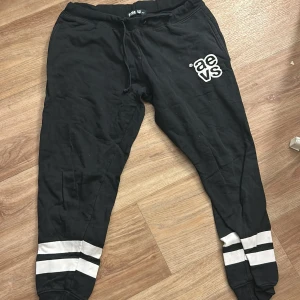 Svarta mjukisbyxor från Svea - Svarta mjukisbyxor från Svea med vita ränder längst ner på benen och tryckt logga på ena låret. Byxorna har dragsko i midjan och är perfekta för en avslappnad stil.