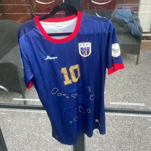 Blå Cabo Verde fotbollströja med nummer 10 - Säljer en blå fotbollströja från Cabo Verde med guldigt nummer 10 på bröstet och ryggen. Tröjan har röda detaljer vid krage och ärmslut, samt vita och guldiga tryck. FCF-logga på bröstet och texten 'NO STRESS' på ryggen.