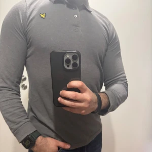 Grå långärmad pikétröja från Lyle & Scott - Säljer en stilren grå långärmad pikétröja från Lyle & Scott med klassisk krage och knappslå. Tröjan har den ikoniska gula loggan broderad på bröstet. Perfekt till jeans eller chinos.