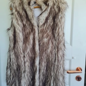 Lång pälsväst från Vera Lucci - Säljer en lång och fluffig pälsväst från Vera Lucci i naturfärgad ton med inslag av brunt, grått och vitt. Västen har en öppen front och är perfekt för att ge din outfit en extra touch. Fodrad insida för extra komfort.