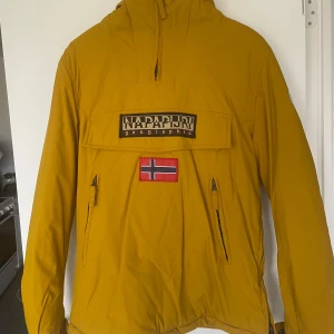 Gul jacka från Napapijri - Säljer en gul jacka från Napapijri med dragkedjor på sidorna och stor ficka framtill med logga och norsk flagga. Jackan har luva och är perfekt för kyliga dagar. 
