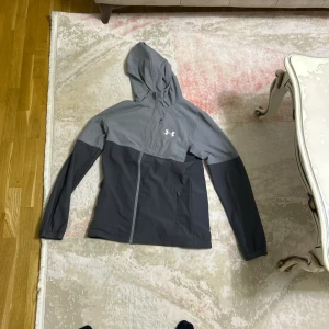 Grå vindjacka med huva från Under Armour - Snygg tvåfärgad grå vindjacka från Under Armour med huva och dragkedja framtill. Jackan har lång ärm och två sidofickor med dragkedja. Perfekt för dig som gillar sportig stil.