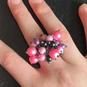 Statementring med färgglada pärlor - Cool ring med stora pärlor i rosa, lila och svart. Pärlorna sitter tätt ihop och ger en unik och lekfull look. Perfekt för dig som vill sticka ut med dina accessoarer!