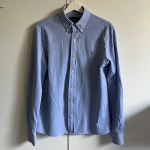 Blå oxfordskjorta från Ralph Lauren - Storlek M men passar S. Klassisk ljusblå oxfordskjorta från Ralph Lauren med button-down krage och broderad logga på bröstet. Skjortan har långa ärmar och vita knappar framtill. Perfekt för en stilren och tidlös look. 