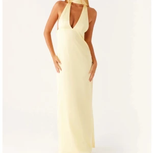 Ljusgul balklänning med halterneck - Säljer en elegant ljusgul långklänning med halterneck och djup v-ringning. Klänningen har öppen rygg med långa band som faller ner baktill och är tillverkad i ett glansigt, mjukt material som ger en lyxig känsla. 