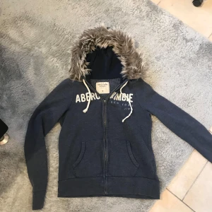 Blå hoodiejacka med fuskpäls från Abercrombie & Fitch - Mysig blå hoodiejacka från Abercrombie & Fitch med dragkedja och vit logga på bröstet. Jackan har huva med fuskpäls och vita snören. Perfekt för kyliga dagar och enkel att matcha till jeans.