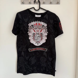 Svart t-shirt från Philipp Plein Junior med tryck - Cool svart t-shirt från Philipp Plein Junior med stort tryck av krona och örn framtill, röda detaljer och logga på ärmar och rygg. T-shirten har korta ärmar och är i mjuk bomull. Perfekt för dig som gillar statement-plagg!                                                        Storlek 16-17
