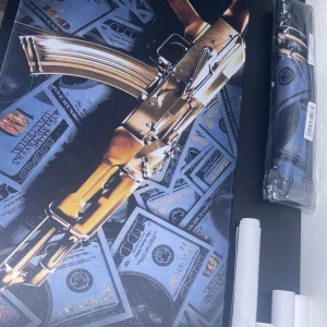 Musmatta med guldfärgad AK-47 och dollarsedlar - Säljer en mussmata med motiv av en guldfärgad AK-47 ovanpå flera amerikanska dollarsedlar. Färgerna är främst guld, blått och svart. Tryckt på canvasduk, levereras rullad. Perfekt för dig som gillar en unik och iögonfallande stil på ditt gamingbord, den är helt ny inte ens öppnad. Tveka inte på att skriva vid minsta fundering! 