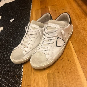 Vita sneakers från Philippe Model - Säljer ett par vita Philippe Model sneakers med svarta detaljer på hälen och grå mocka framtill. Skorna har snörning och en klassisk låg modell med rund tå. Perfekta för en stilren vardagslook.
