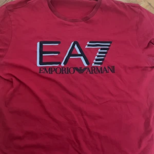 Röd t-shirt från Emporio Armani EA7 - Snygg röd t-shirt från Emporio Armani EA7 med stor logga framtill. Klassisk rund halsringning och korta ärmar. Perfekt för dig som gillar stilrena märkesplagg med tydlig branding.