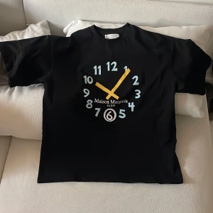 Svart t-shirt från Maison Margiela - Cool svart t-shirt från Maison Margiela med stort klockmotiv och tryckt logga på bröstet. T-shirten har rund hals och korta samt breda ärmar. Perfekt för dig som gillar unika designer och streetwear.