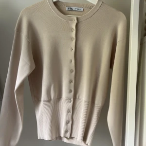 Oanvänd beige kofta från Zara🥰 - Säljer en stilren beige kofta från Zara med rund halsringning och knappar framtill. Tröjan har långa ärmar och ribbad nederkant som ger en snygg passform. Perfekt att styla till olika outfits!