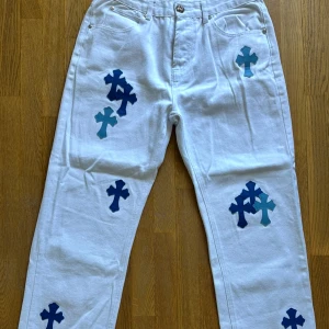 Vita Levis 550 jeans med blå kors. Storlek 32. - Unika vita Levis 550 jeans med flera blå korsapplikationer på benen. Klassisk femficksmodell med silverfärgade knappar och Levis-märke bak. Rak passform och tillverkade i kraftigt jeanstyg. 