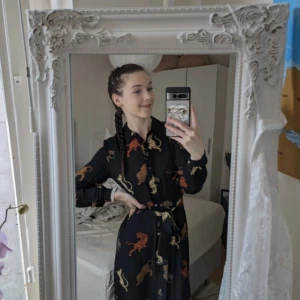 Svart långklänning med lejonmönster från ONLY - Säljer en svart långklänning från ONLY med färgglada lejonmönster i rött, beige och gult. Klänningen har lång ärm, krage och knappar framtill samt ett matchande bälte i midjan. Perfekt för dig som vill sticka ut med ett unikt print!