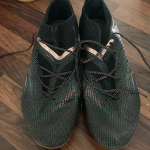 Svarta Puma fotbollsskor med snörning - Säljer ett par svarta Puma fotbollsskor med högt skaft och snörning. Skorna har mönstrad ovandel och dobbar för spel på gräs. Snygga detaljer i kopparfärg och svart. Perfekta för dig som vill ha bra grepp på planen.