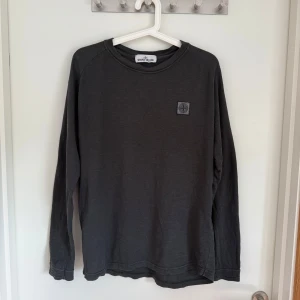 Mörkgrå långärmad tröja från Stone Island - Säljer en stilren mörkgrå långärmad tröja från Stone Island med klassisk logotyp på bröstet. Tröjan har en enkel design och rund halsringning, perfekt för en avslappnad look.