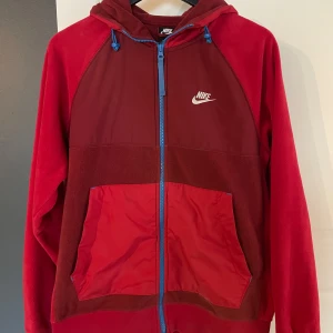 Röd fleecejacka med huva från Nike - Röd fleecejacka från Nike med huva och blå dragkedja. Jackan har två stora fickor framtill och Nike-logga på bröstet. Det är storlek S på jackan och är i bra skick.