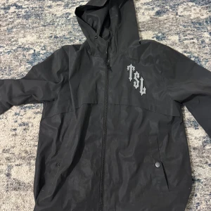 Trapstar jacka - Säljer en svart vindjacka från Trapstar London med huva och dragkedja. Jackan har tryck med 'TSL' på bröstet och stort 'Trapstar London Shooters' tryck på ryggen. Perfekt för streetwear-stil och enkel att matcha.
