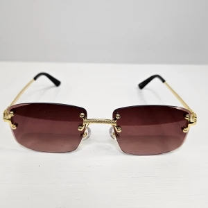 Guldiga solglasögon med bruna glas - Stilrena solglasögon med rektangulära, bruna glas och tunna guldiga bågar. Glasögonen har svarta ändar på skalmarna och en elegant detalj vid näsbryggan. Perfekta för dig som vill ha en trendig accessoar med lyxig känsla.