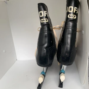 Svarta skridskor från JOFA med snörning - Klassiska svarta skridskor från JOFA med vit snörning och bruna detaljer vid skaftet. Skorna har rund tå och är tillverkade i skinn med robust platt sula och stålskena. Skydd är inkluderade i priset. Perfekta för vinterns isaktiviteter.