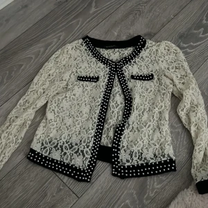 Vit spetskofta med pärldetaljer från Red London - Säljer en vit spetsjacka från Red London med svarta kanter och pärldetaljer. Kofta har långärmade spetsärmar och dekorativa fickor framtill. Perfekt för dig som vill sticka ut med en unik och elegant stil.