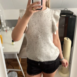 Stickad ärmlös tröja med volang från Saint - Superfin ljusgrå stickad tröja från Saint med volangdetaljer på axlarna. Ärmlös modell med rund halsringning och diskret melerat mönster. Perfekt att styla med jeans eller kjol för en chill och trendig look. Knappt använd.