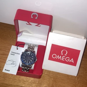 Omega Seamaster klocka - Snygg Omega Seamaster med blå urtavla och silverfärgat armband i metall. Klockan har vågigt mönster på urtavlan, tydliga index och datumfönster. Kommer i original röd Omega-box med tillhörande certifikat och etikett. Perfekt för dig som gillar klassisk och sportig stil.