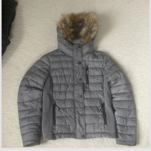 Grå quiltad dunjacka med fuskpäls - En vinterjacka från superdry, dne är i fint skick men kommer inte till användning längre. Den är i storlek L men det är barnstorlekar så den är XS/S i ”vanlig” storlek tror jag. 