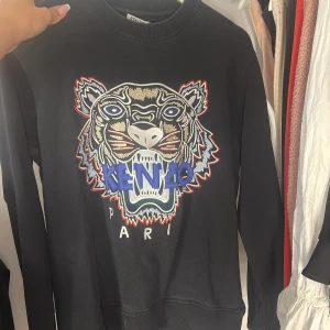 Svart Kenzo sweatshirt med tigertryck - Svart sweatshirt från Kenzo med ikoniskt broderat tigerhuvud och texten 'KENZO PARIS' framtill. Tröjan har rund hals, långa ärmar och ribbade muddar. Perfekt statement-piece för dig som gillar streetstyle och vill sticka ut.