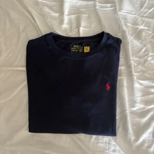 Mörkblå t-shirt från Polo Ralph Lauren - Snygg mörkblå t-shirt från Polo Ralph Lauren med klassisk röd broderad logga på bröstet. T-shirten har rund halsringning och är tillverkad i mjuk bomull. Perfekt för en clean och stilren look. Den är nästan aldrig använd och är precis som ny.