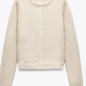 Beige stickad tröja med lång ärm - Mysig beige stickad tröja med rund halsringning och lång ärm. Tröjan har en enkel design med en diskret söm framtill och ribbade muddar vid ärmslut och nederkant. Perfekt för lager-på-lager under kyliga dagar.