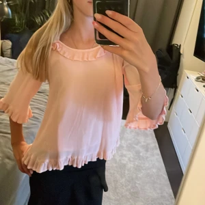 Ljusrosa blus med volang från Lindex - Supersöt ljusrosa blus från Lindex med volangdetaljer vid halsringning, ärmslut och nederkant. Blusen har offshoulder-design och trekvartsärm, vilket ger en luftig och trendig look. Perfekt för dig som gillar romantiska och feminina plagg.🥰🥰