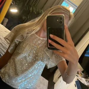 Glittrig topp med volang från Lindex - Superfin glittrig topp från Lindex med korta volangärmar och rund hals. Toppens tyg är täckt av silvriga paljetter som ger ett riktigt festligt intryck. Perfekt för dig som vill sticka ut och glänsa! Luftig passform och knäppning bak i nacken.