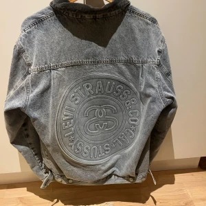 Levi's x Stüssy jeansjacka med tryck dubbelsidig - Unik jeansjacka från Levi's och Stüssy med stort broderat logomärke på ryggen och coolt Stüssy-tryck med 8-ball och ränder i rött och blått. Jackan har svarta quiltade ärmar, klassisk krage och knappar framtill. Perfekt statement-plagg för streetwear. Kan bäras på båda hållen dubbel disig alltså 2jackor
