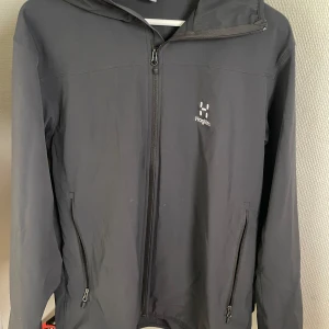 Svart vindjacka från Haglöfs med huva - Snygg svart vindjacka från Haglöfs med minimalistisk design och praktisk huva. Jackan har dragkedja framtill, två sidofickor med dragkedja och diskret logga på bröstet. 