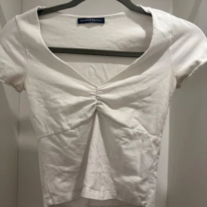 Vit topp med rynk från Brandy Melville - Supersöt vit topp från Brandy Melville med korta ärmar och v-ringning. Toppen har snygg rynkdetalj framtill och är tillverkad i mjuk bomull. Perfekt att styla med!