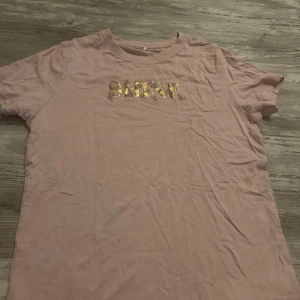Rosa t-shirt med guldtext från Name It - Söt rosa t-shirt från Name It med korta ärmar och rund hals. Framtill står det 'SHINE' i glansig guldtext med små broderade blommor runt bokstäverna. Perfekt för dig som gillar en mjuk och bekväm stil med lite extra bling.