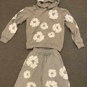 Denim Tears hoodie - Säljer en grå hoodie med stora vita blommönster över hela tröjan. Tröjan har huva, långa ärmar och ribbade muddar. Perfekt för dig som gillar en chill och trendig stil med lite extra detaljer.