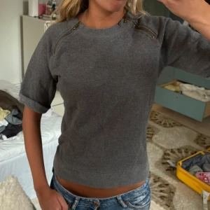 Grå kortärmad sweatshirt med dragkedjor - Grå sweatshirt i mjuk bomull med korta ärmar och coola dragkedjor vid axlarna. Tröjan har en avslappnad passform och rund halsringning. 