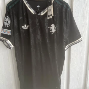 Svart Adidas fotbollströja Yildiz 10 - Snygg svart fotbollströja från Adidas med Yildiz och nummer 10 i guld på ryggen. Tröjan har korta ärmar, vita detaljer vid krage och ärmslut samt diskret blommönster i tyget. Broderad Adidas-logga och lejonmärke på bröstet, samt UEFA Foundation-märke på ärmen.