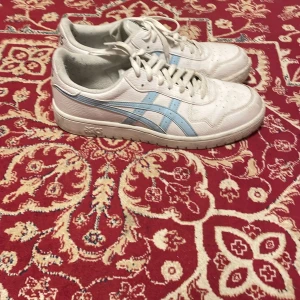Asics Japan  - Snygga vita Asics sneakers med klassiska ljusblå ränder på sidorna. Skorna har snörning, rund tå och platt sula. Tillverkade i skinn med perforerade detaljer upptill för extra ventilation. Perfekta för dig som gillar en clean och sportig look.