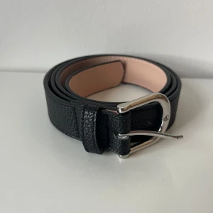 Svart skärp med silverfärgat spänne - Stilrent svart bälte i skinn med klassisk struktur och ett snyggt, silverfärgat metallspänne. Strl M. Flera hål för att kunna reglera längden. Bredd: 3,5cm. Längd 108cm.