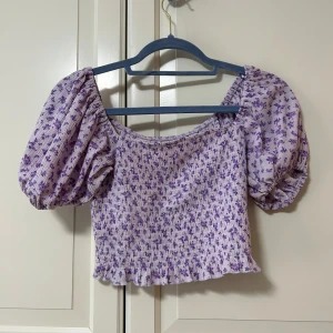 Topp från Gina Tricot - Supersöt lila blommig topp från Gina Tricot med puffärmar och smockad, croppad passform. Toppen har volangkant nertill. Super snygg och perfekt för sommar och utekvällar! Använd typ en gång 
