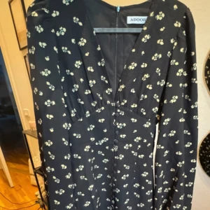 Svart blommig långklänning från Adoore - Supersnygg svart långklänning från Adoore med små vita blommor, markerad midja och v-ringning. Klänningen har långa puffärmar och knappar längs hela framsidan. Perfekt för dig som gillar romantisk och trendig stil.