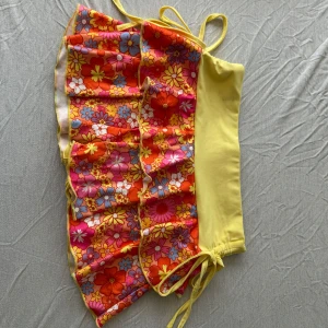 Gul och blommig bikini med volang - Superfin bikini med blommigt mönster i orange, rosa, blått och vitt samt enfärgade gula partier. Toppen har smala axelband och volangdetaljer framtill. Bikinin har knyt i sidorna och är tillverkad i ett stretchigt syntetmaterial, perfekt för stranden.