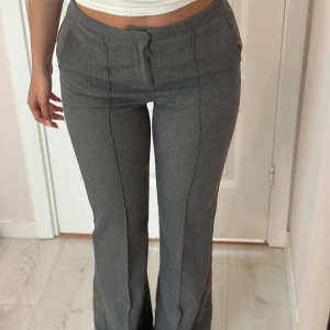Grå kostymbyxor med bootcut - Snygga grå kostymbyxor med bootcut och pressveck längs benen. Byxorna har hög midja, diskreta fickor fram och bak samt dragkedja och knapp framtill. Perfekta för en clean och stilren look.