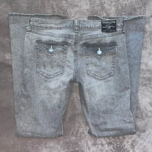 True Religion Joey grå jeans, bootcut - Grå jeans från True Religion, modell Joey med låg midja och bootcut. Snygga detaljer på bakfickorna med kontrastsömmar och logoknappar. Jeansen har klassisk femficksdesign och är tillverkade i mjukt denimtyg.