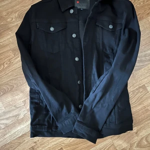 Svart jeansjacka med knappar - Snygg svart jeansjacka med klassisk design och silverfärgade knappar framtill. Jackan har två bröstfickor med lock och knappar samt raka ärmar. Perfekt för dig som vill ha en stilren och tidlös look.