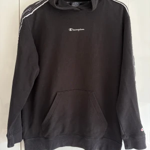 Svart Champion hoodie med logga - Svart hoodie från Champion i storlek XS. Lite oversized i modellen, så den är lite stor för xs. Klassisk modell med huva, magficka och broderad Champion-logga på bröstet. Broderat band med logga längs axlarna. 