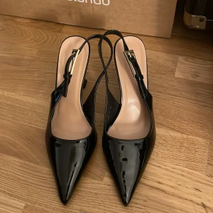 Svarta lackpumps med spetsig tå - Snygga svarta pumps i lack med spetsig tå och öppen häl. Skorna har en elegant hög klack och justerbart spänne i guld. Perfekta för dig som vill sticka ut med en stilren och trendig look. Skorna är gjorda för plus side kvinnor oxå jätte bekväma har andvänt dom en gång bara 